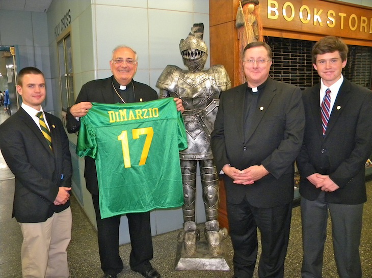 Bishop Nicholas DiMarzio celebrated Mass on Halloween at Holy Cross H.S., Flushing, marking the 17th anniversary of his ordination as a bishop. The school community presented him with a personalized Knights’ athletic jersey to mark the occasion. Pictured are, from left, senior Brian Lloyd; Bishop DiMarzio; Father Walter Jenkins, C.S.C., president; and senior Robert Bowe. Photo © Jim Mancari