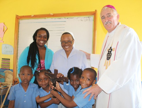 Bishop Tiedemann with teacher Stephanie Watson, Sister Susan N’gendo, A.S.N., and students of St. Theresa Kindergarten School with a crucifix for a newly constructed classroom.