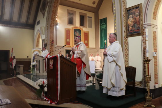 Auxiliary Bishop Witold Mroziewski was the main celebrant at the jubilee Mass. He commended the community for preserving the Polish culture, spirit and language through works at the parish and the school. He spoke at the lectern adorned with the Polish national colors and the Polish national emblem. Behind him is the American flag and above him is a depiction of the Risen Lord. 